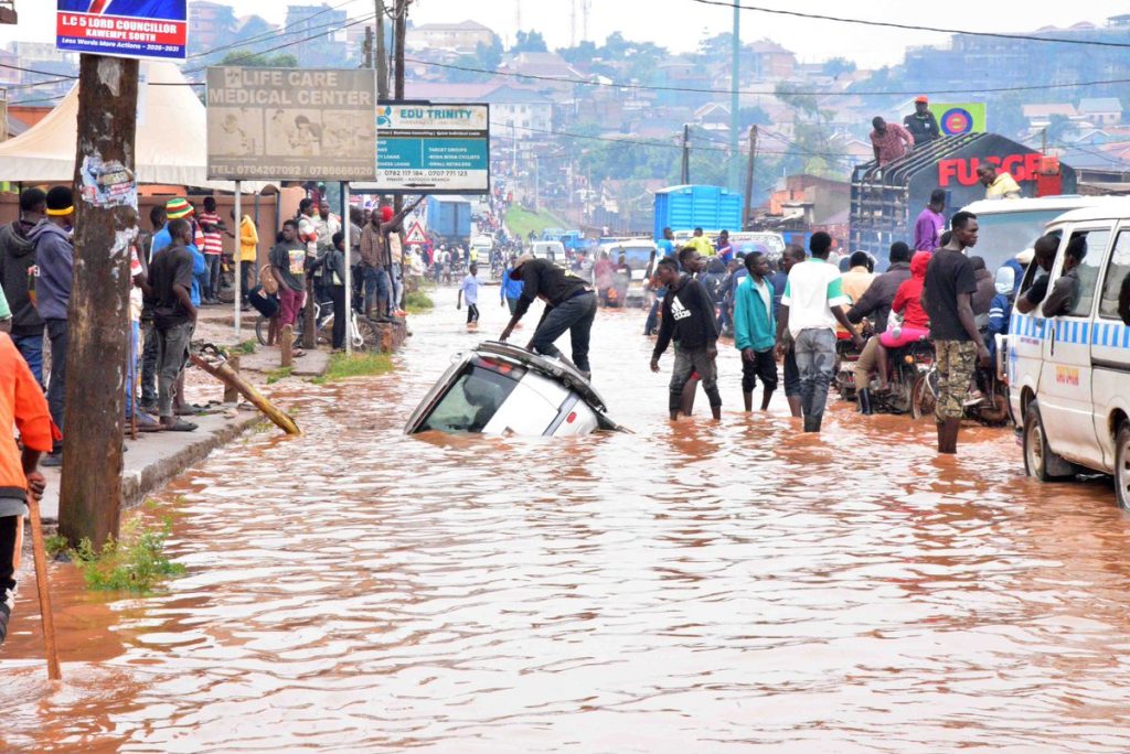 Flooded street in Kampala with vehicles and people wading through water after heavy rainfall – highlighting poor drainage and urban planning challenges in Uganda.